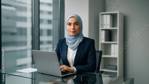 Portrait of a professional Muslim woman wearing a tailored navy blazer, light blue hijab wrapped neatly, and white blouse, sitting at a modern glass desk, laptop open, looking into the camera.