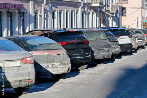 Dirty salty cars tightly parked in diagonal parking spaces on narrow city street with visible mud stains and salt residue on vehicle bodies during sunny winter day. Vehicles covered in salt and dirt