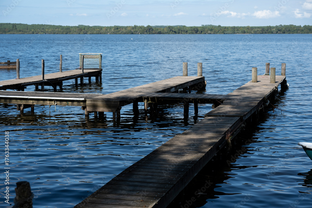 Fototapeta premium The wooden piers at Lac de Leon stretch into the calm blue waters. Small boats are moored nearby, creating a peaceful atmosphere surrounded by lush trees and clear skies.
