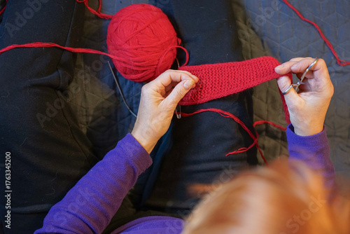 Woman knitting red wool fabric, top view. Ball of yarn, needles, and cozy home atmosphere.