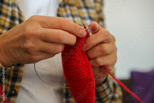 Woman in checkered shirt knitting red fabric with needles, close-up of hands and yarn. Cozy home hobby and creativity.