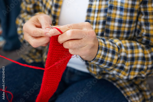 Woman in checkered shirt knitting red fabric with needles, close-up of hands and yarn. Cozy home hobby and creativity.