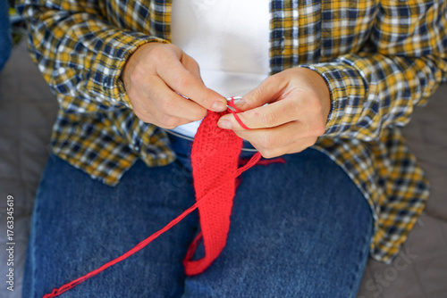 Woman in checkered shirt knitting red fabric with needles, close-up of hands and yarn. Cozy home hobby and creativity.