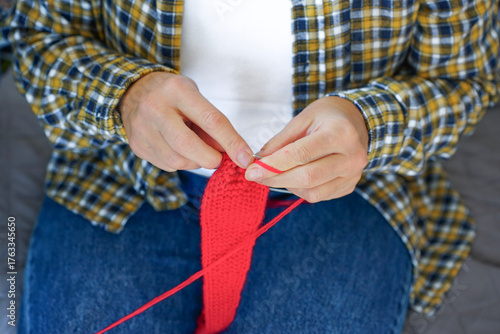 Woman in checkered shirt knitting red fabric with needles, close-up of hands and yarn. Cozy home hobby and creativity.