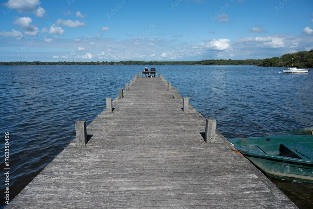 Fototapeta premium An elderly couple sits side by side on a bench at Lac de Leon. They enjoy the calm waters and scenic beauty of the Landes region on a clear day.