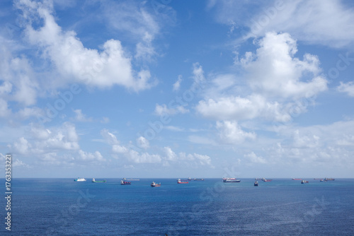 Panoramic view of multiple cargo ships and tankers anchored in the calm blue sea under a bright sky with scattered clouds.