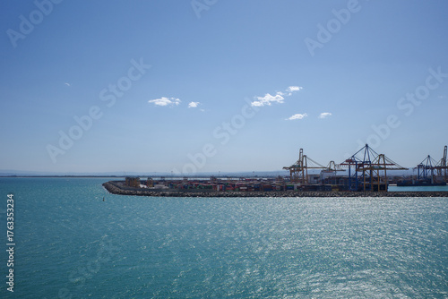 Industrial container port with large cargo cranes on a sunny day, viewed from the sea.