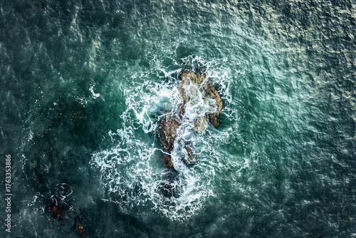Abstract aerial close-up of lone submerged rock in turbulent textured turquoise and grey coastal waters in Ireland, creating bright white foam