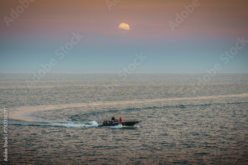 Fast black powerboat crossing Irish Sea under full moon at twilight, two people aboard in dramatic seascape.