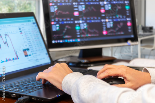 Hands typing on laptop while checking stock market trends on multiple screens in a modern office setup