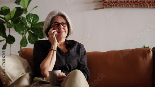 Happy retired woman relaxing on comfortable sofa, talking on smartphone and drinking tea