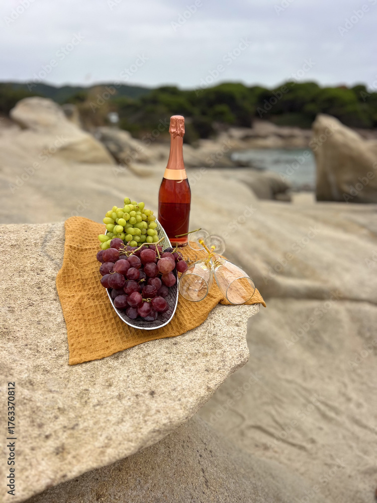 Naklejka premium A bottle of wine with a bunch of grapes on a sandy beach, captured in soft natural light. Summery coastal mood, relaxed vacation atmosphere, elegant still life outdoors. 