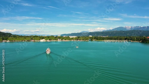 Aerial landscape of Worthersee in Klagenfurt, Austria featuring charming waterfront homes, lush greenery, tourist boat and serene blue waters under a clear sky.