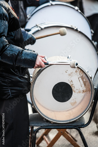 Drummer in black puffer jacket plays bass drum with mallets outdoors — close-up of hands, worn drum head, and winter performance energy perfect for music, culture or event themes.