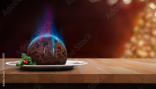 A low angle view of a traditional Christmas pudding with a blue flame of lit brandy on a round plate with a sprig of holly, berries on a wooden table against a dark festive background.