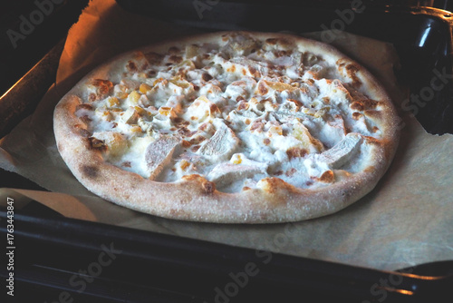 Pizza with pineapple and chicken pieces on a baking sheet on parchment in the oven. Hawaiian pizza on a black background. Cooking at home, the theme of recipes and nutrition