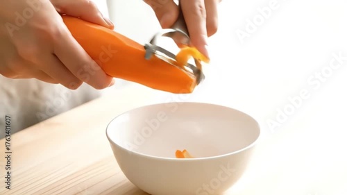 Close-up of hands peeling a fresh orange carrot with a vegetable peeler over a white bowl preparing healthy ingredients for cooking a nutritious meal in a brigh