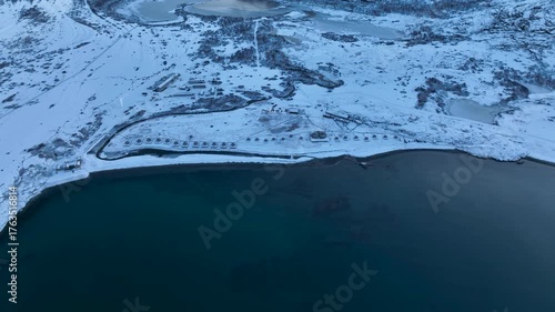 Aerial view of a remote arctic landscape with a row of small domes or tents along the coastline, snow-covered hills, and frozen lakes inland; a small part of a computer screen interface is visible on 