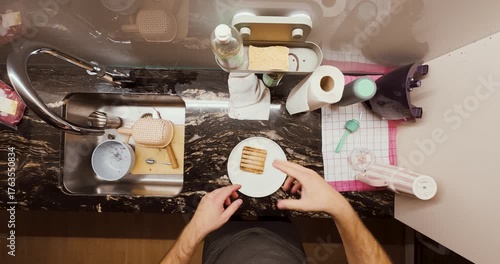 A man prepares a sandwich in his kitchen and accidentally burns his fingers on the hot toast. The scene captures a relatable everyday kitchen mishap from a top-down perspective.