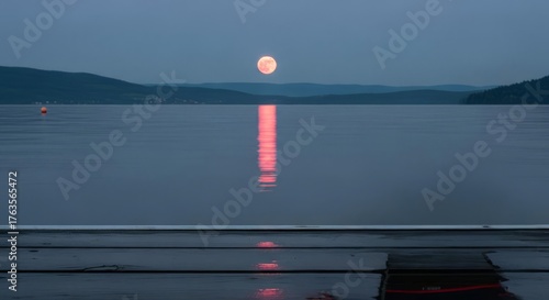 Serene full moon reflecting a pink glow on a tranquil lake with distant mountains at dusk or night, peaceful natural landscape.