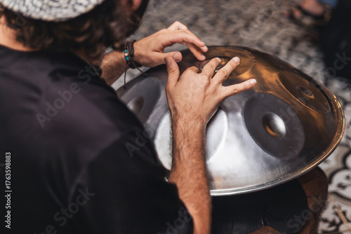 A young man with curly hair plays a handpan instrument. He wears a black shirt and is seated on a patterned floor. The scene captures a moment of musical creativity.