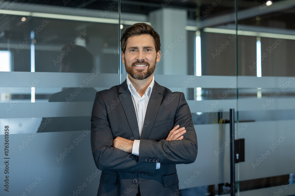 Obraz premium Businessman with a beard and dark suit standing confidently with arms crossed in a modern office building, looking directly at the camera with a friendly smile