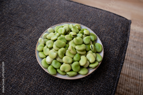 Photos High resolution image of a ceramic bowl filled with fresh green fava beans placed on a textured dark fabric surface