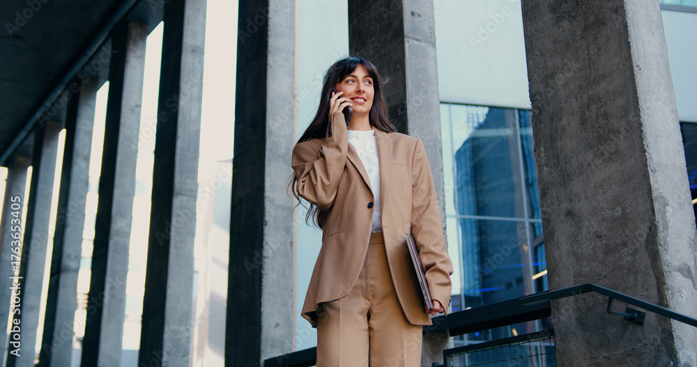 Fototapeta premium Confident female professional standing outside a modern office building, holding a laptop in one hand and speaking on the phone with a colleague, engaged in a serious business conversation