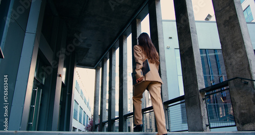Rear view of confident businesswoman with long flowing hair, wearing tailored formal suit, walking up outdoor stairs while holding laptop in one hand, approaching modern office building