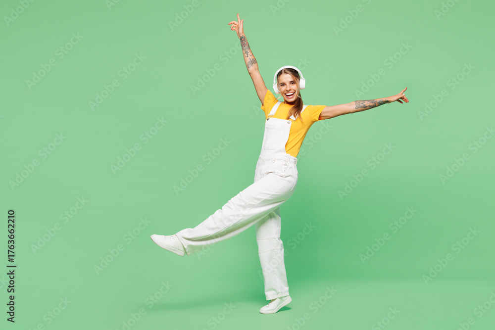 Fototapeta premium Full body young happy woman she wear yellow t-shirt overall casual clothes listen to music in headphones raise up hands dance isolated on plain pastel light green background studio. Lifestyle concept.