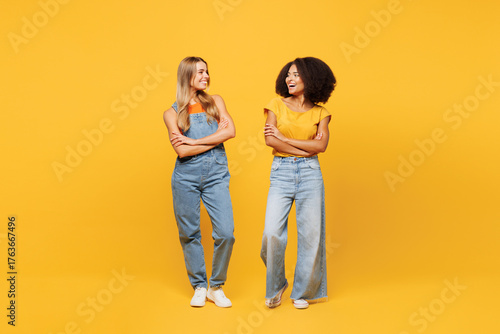 Obraz na plátne Full body two young friends buddies women wearing denim casual clothes together stand look to each other talk speak smile isolated on plain yellow orange background studio portrait