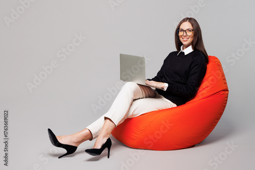 Full body young smart employee IT business woman corporate lawyer wear black clothes glasses work in office sit in bag chair hold use work on laptop pc computer isolated on plain grey color background