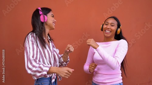 Happy young latin women dancing together in the city using wireless headphones - Technology, generation z and urban lifestyle concept 