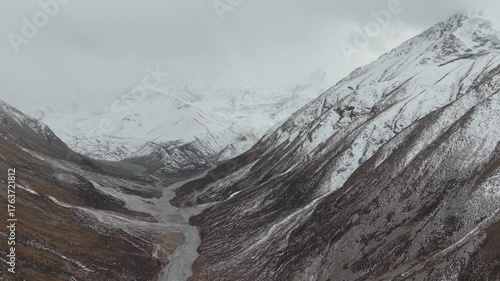 Aerial view captures the majestic snow-capped mountains surrounding a winding river in a deep valley, showcasing the natural beauty and rugged terrain, camera pans to reveal the expansive landscape