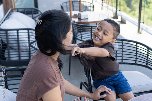 A little boy playfully pulling his mother’s hair while laughing together on a bright and windy restaurant balcony.