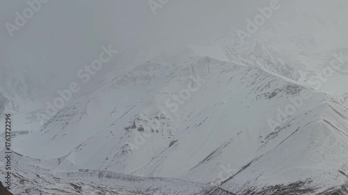 Snowy mountain landscape reveals intricate textures and patterns of rugged terrain, as the camera gradually zooms in, showcasing the dramatic contrasts of snow and rock formations