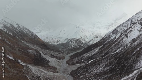 Aerial perspective captures snow-capped mountains framing a winding river flowing through a rugged valley, showcasing the dramatic landscape and atmospheric conditions, camera slowly zooms out