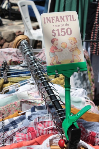 Roupas infantis com cabides em arara. Etiqueta promocional onde diz: infantil a partir de 150, feira ao ar livre, second hand e moda circular