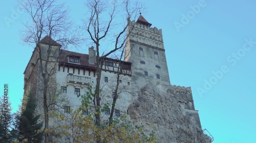 Historic Bran Castle, Romania low angle day exterior, perched on a rocky cliff surrounded by trees under a clear blue sky.