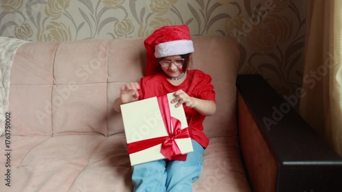 A girl dressed in a red T-shirt and a Santa Claus hat opens a New Year's gift. Christmas. A gift for Christmas