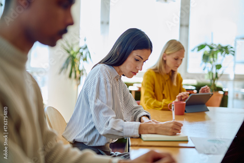 Valokuva Young woman taking notes while collaborating with diverse colleagues in a modern