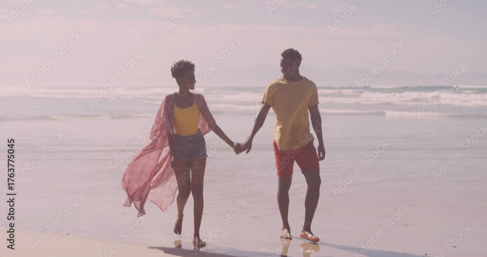 Naklejka premium Walking barefoot couple wearing pink shawl, tank top, swim shorts on wet sand with gentle waves