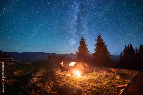 Hiker woman relaxes by campfire, illuminated by its warm glow, with tent nearby under stunning, star-filled sky. Milky Way stretches overhead, adding magical touch to serene mountain setting.