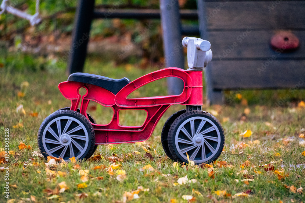 Fototapeta premium Red plastic toy bike on autumn grass – nostalgic childhood scene with fallen leaves, symbol of play, innocence, and fleeting moments of youth