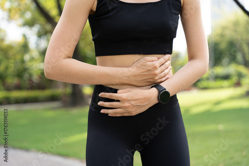 Young woman clutching stomach outdoors in pain, possibly suffering from abdominal cramps or digestive issues during exercise. Concept of health problems, stomach ache, fitness discomfort.