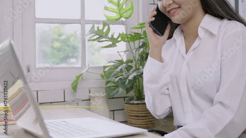 Closeup of happy young Asian female teacher sitting smiling and typing keyboard on laptop computer and using her mobile phone technology talking at her desk in classroom. Back to school concept. 
