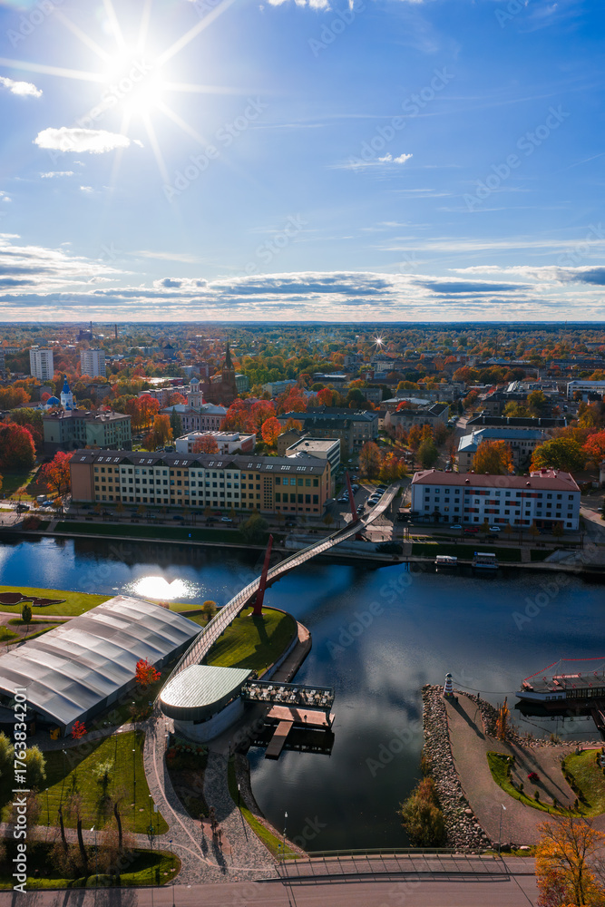 Obraz premium Aerial view shows Jelgava, Latvia in late afternoon. The Driksa River flows under a pedestrian bridge, with a riverside promenade, pavilion, and fall trees with reds and golds.