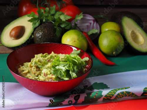 Guacamole in a red bowl, with ingredients at the back. All displayed over a Mexican flag.