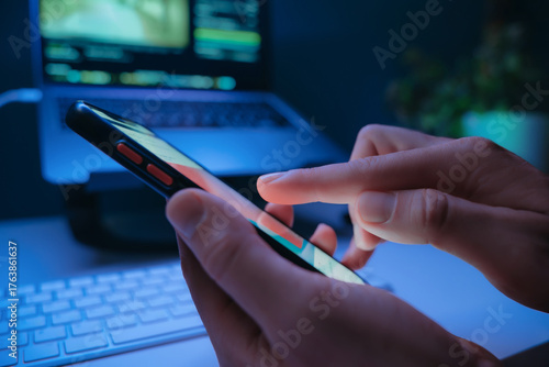 Close-up of persons hand taping on smartphone screen with social media feed at night with laptop on background. Digital addiction and modern online habits. 