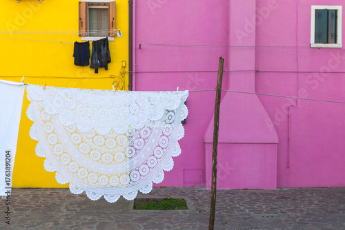 A large, intricately crocheted white tablecloth hangs on a clothesline, drying against vibrant pink and yellow house walls. A concept of traditional craftsmanship and daily life in a colorful village.